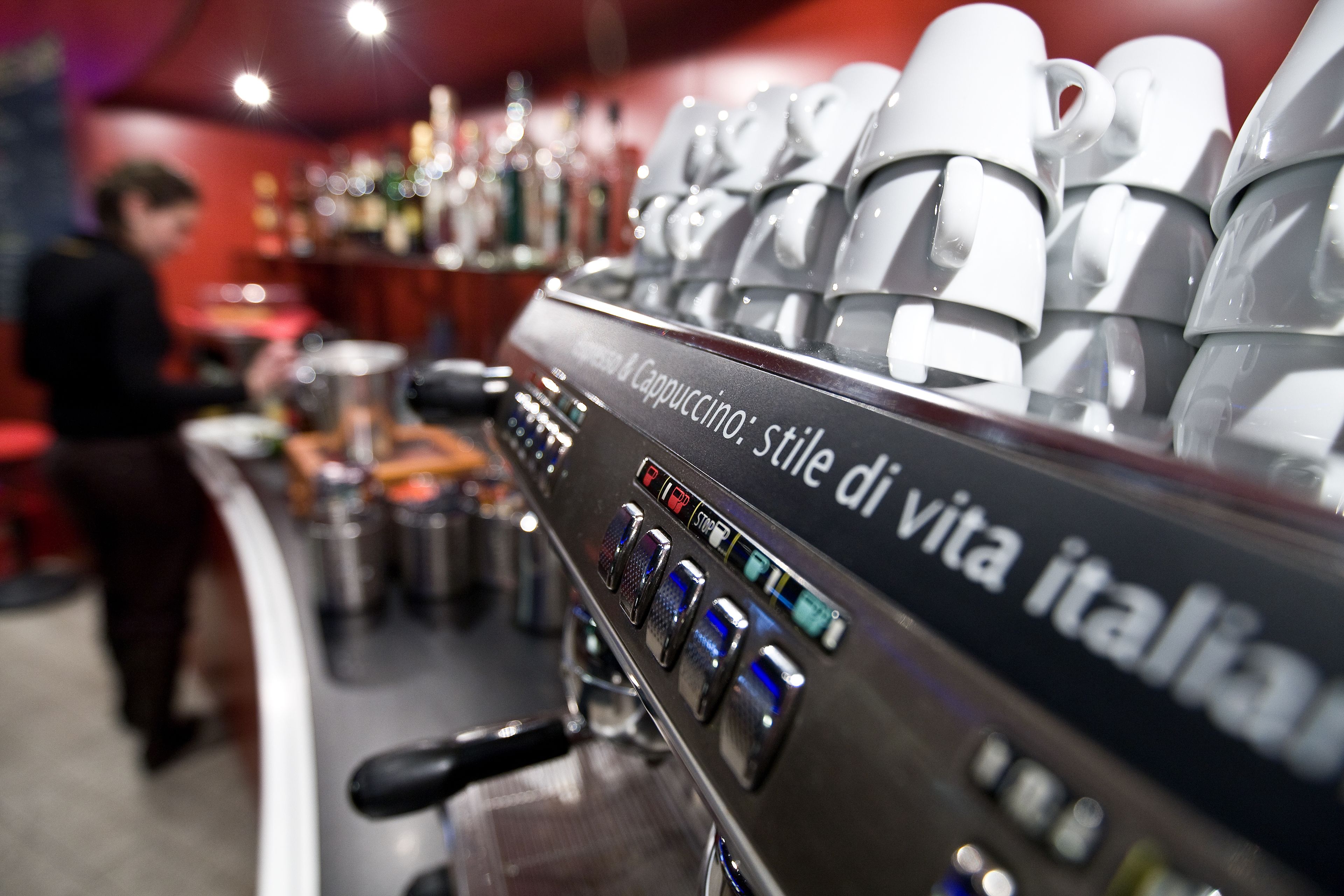 A coffee machine with multiple coffee cups stacked above it, and a person working behind the counter.