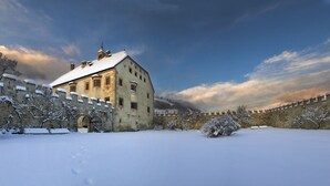 A historic building with a snow-covered roof and walls, surrounded by a stone wall and snow-covered trees.