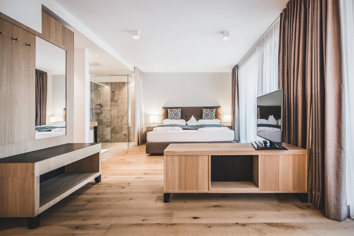 Une chambre d’hôtel moderne avec un plancher en bois, un grand lit avec une tête de lit, une table de chevet, un miroir et un espace douche.