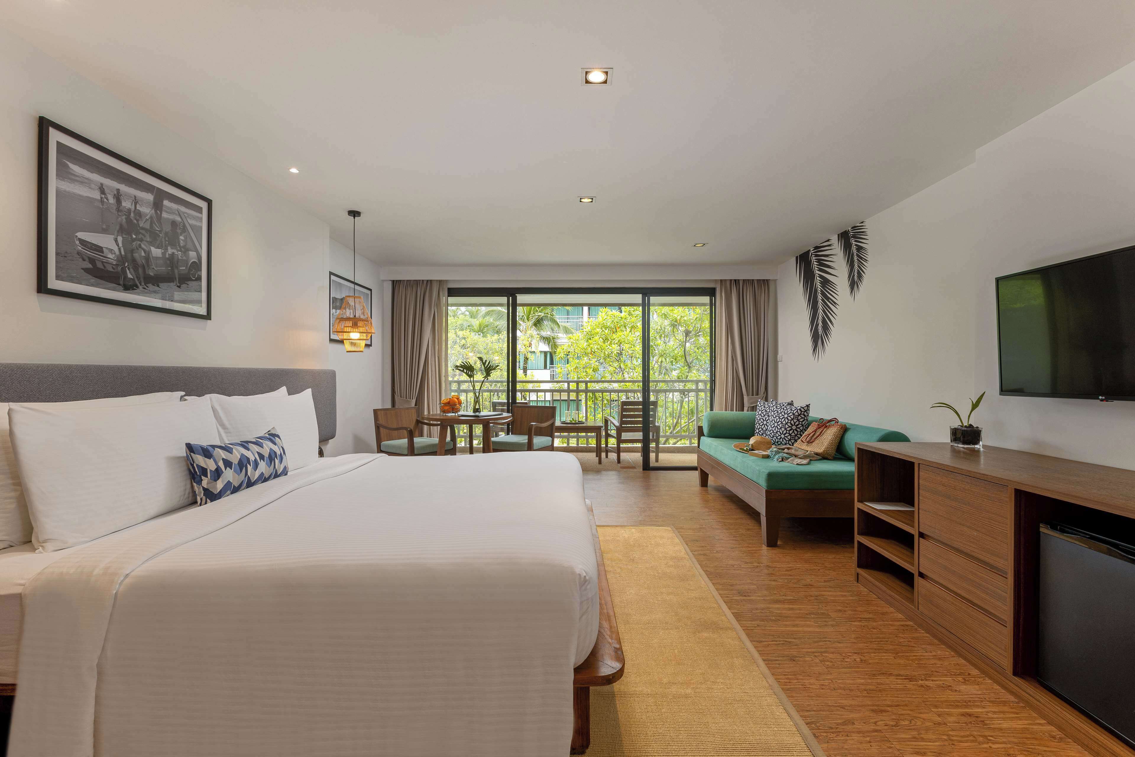 A hotel room with a large bed, a desk, a sofa, a TV, and a balcony with a view of greenery.