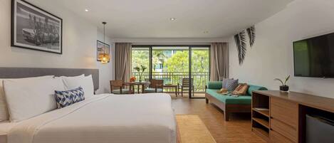 A hotel room with a large bed, a desk, a sofa, a TV, and a balcony with a view of greenery.