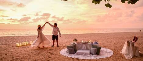 Une cérémonie de mariage organisée sur la plage, avec une table et des chaises, une pancarte indiquant « JOYEUX ANNIVERSAIRE » et un couple qui danse.