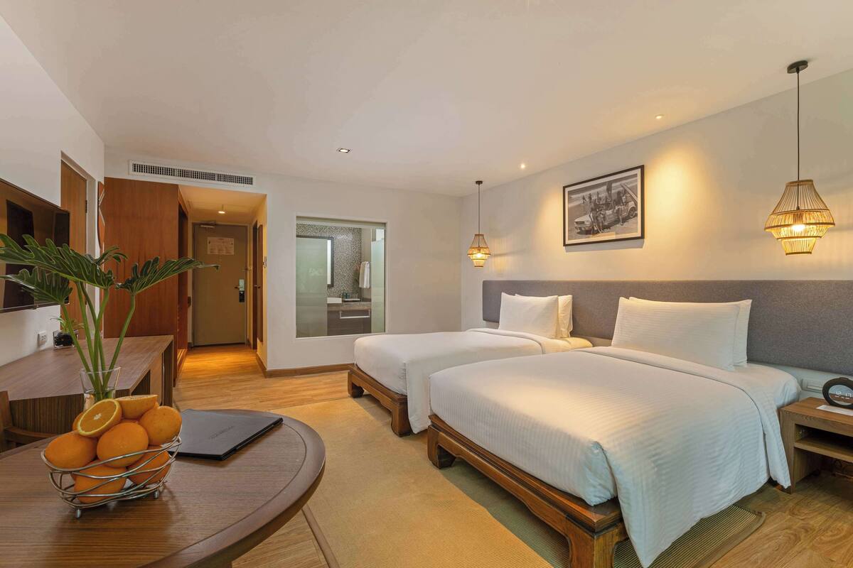 Une chambre d’hôtel avec deux lits, une table à manger agrémentée d’un panier de fruits, un ordinateur portable et un tableau accroché au mur.
