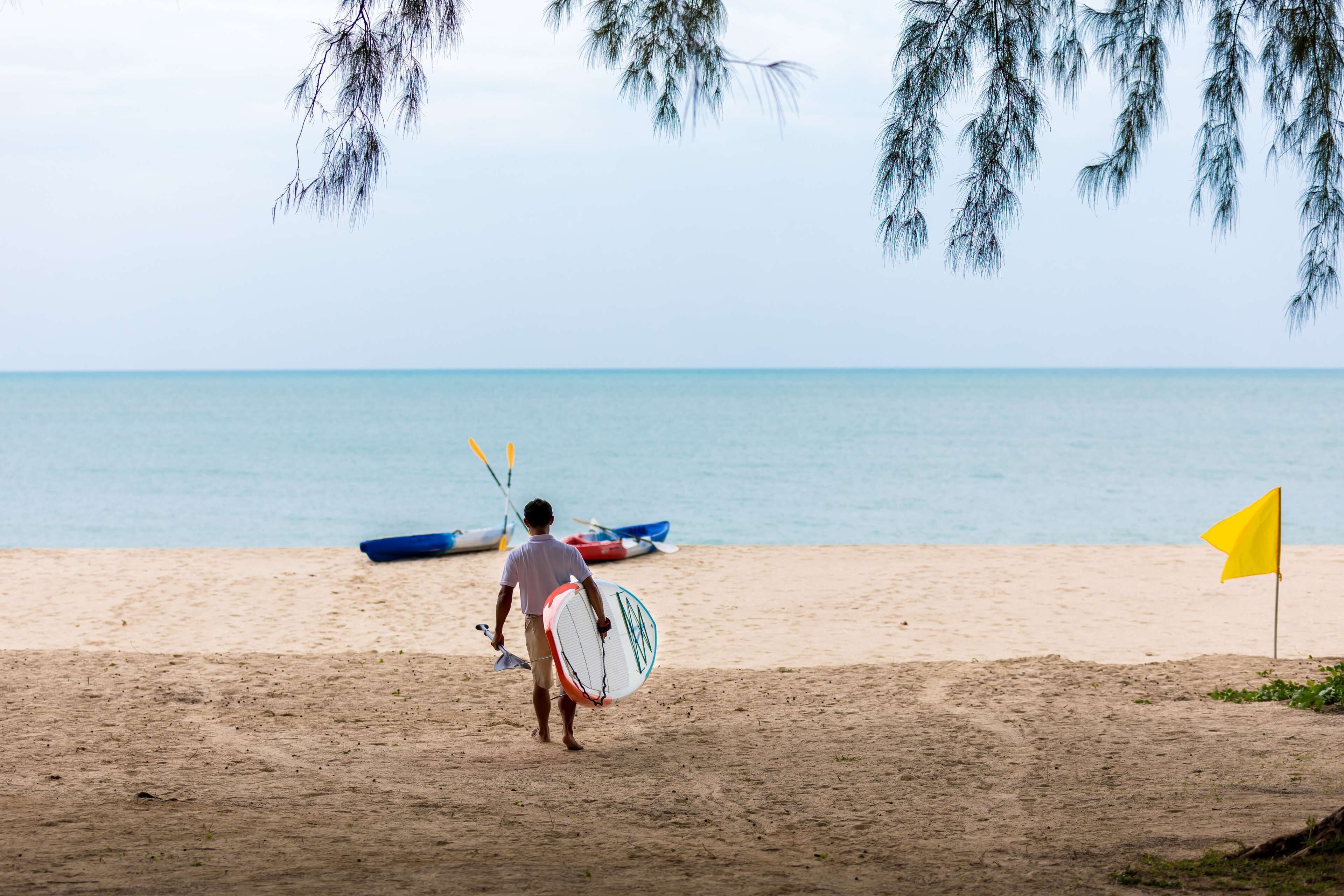 A beach scene with a person carrying a surfboard, a flag, and a boat in the background.