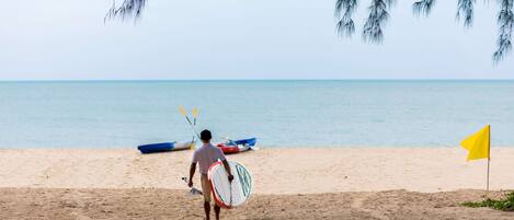 A beach scene with a person carrying a surfboard, a flag, and a boat in the background.
