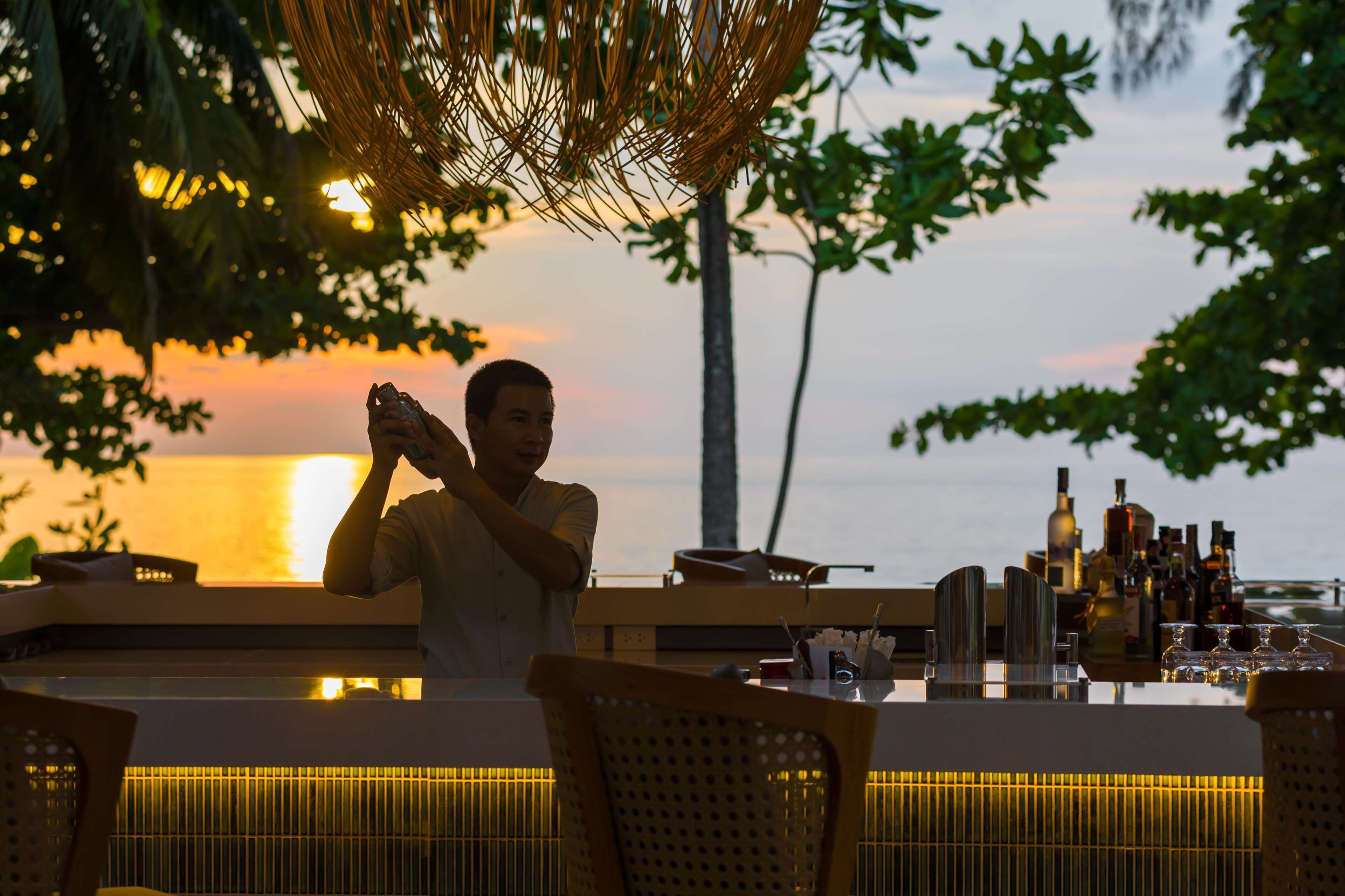 Uma pessoa fotografando em um bar com vista para o pôr do sol sobre a água.