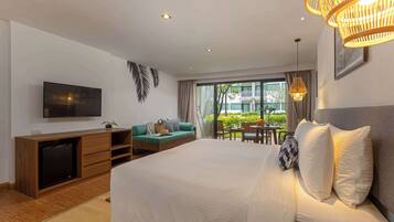 A hotel room with a large bed, a TV, a sofa, a dining table, and a view of greenery outside.