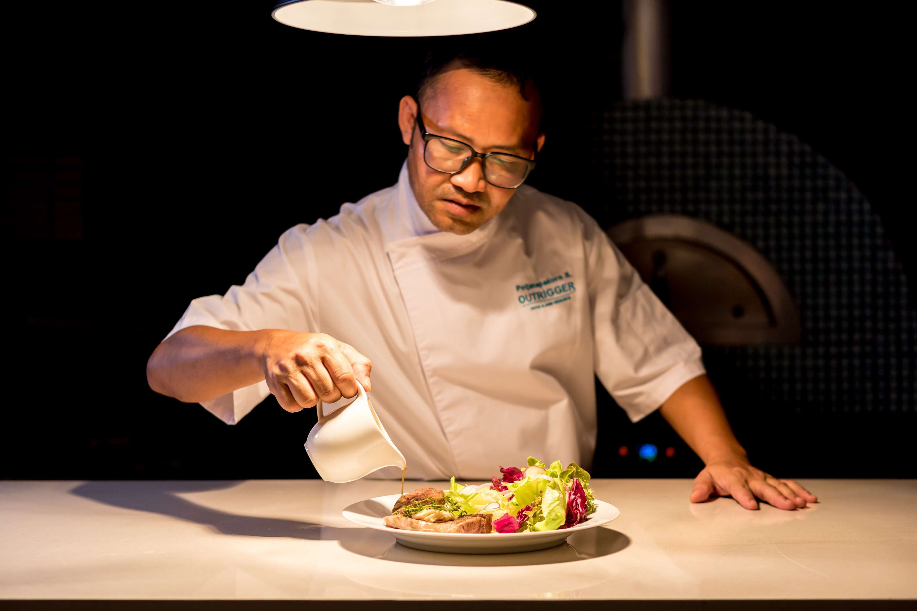 A chef in a white uniform pouring sauce onto a plate of food.