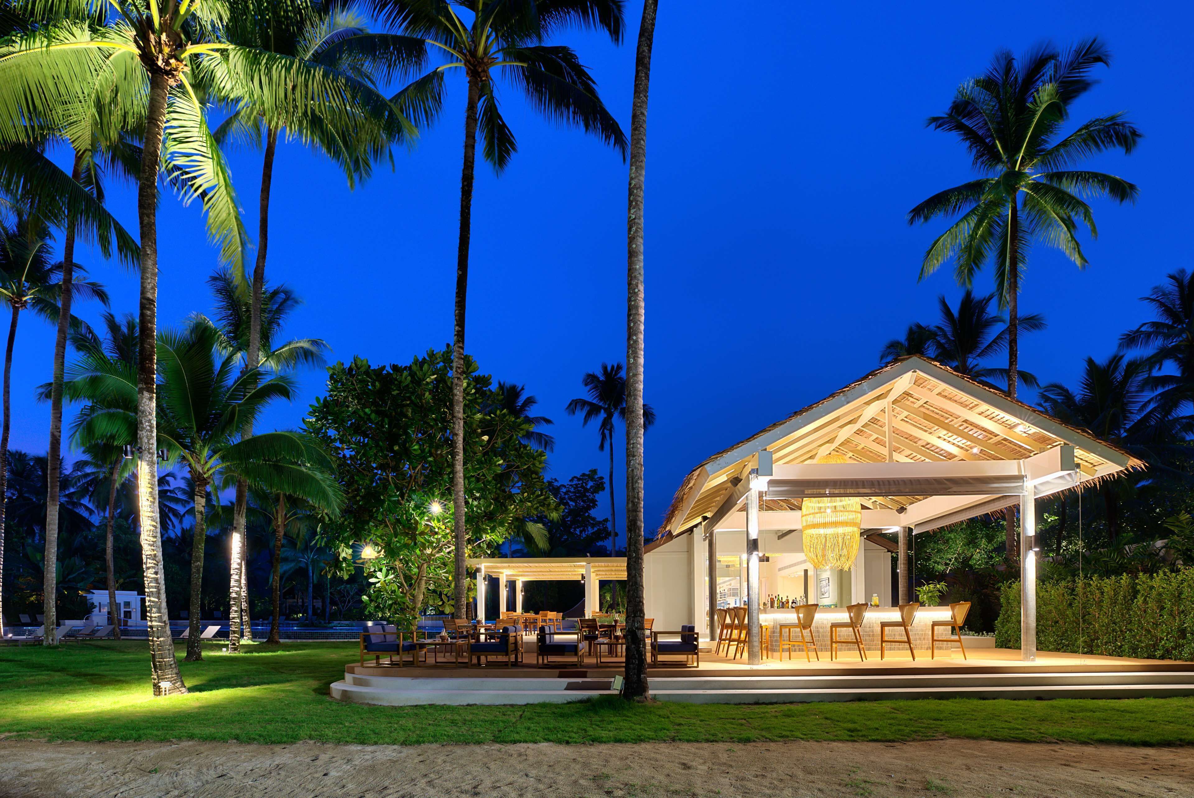 A resort with a covered outdoor seating area, palm trees, and a well-lit interior.