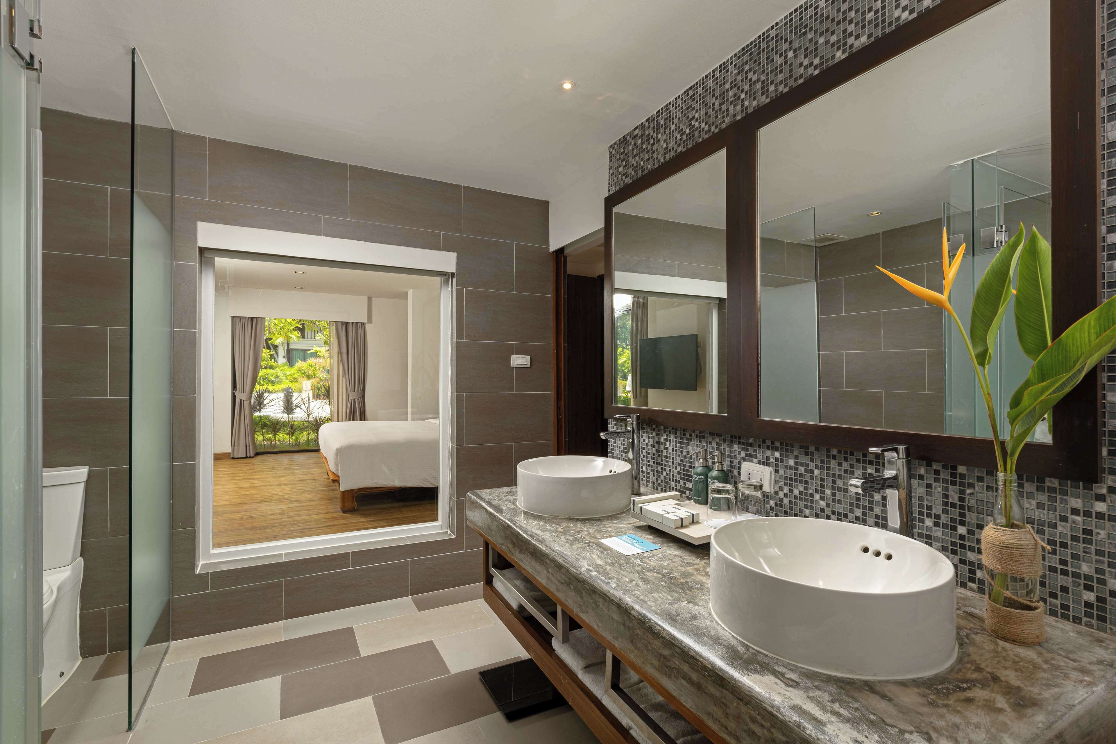 A modern bathroom with two sinks, a large mirror, and a view into the bedroom.