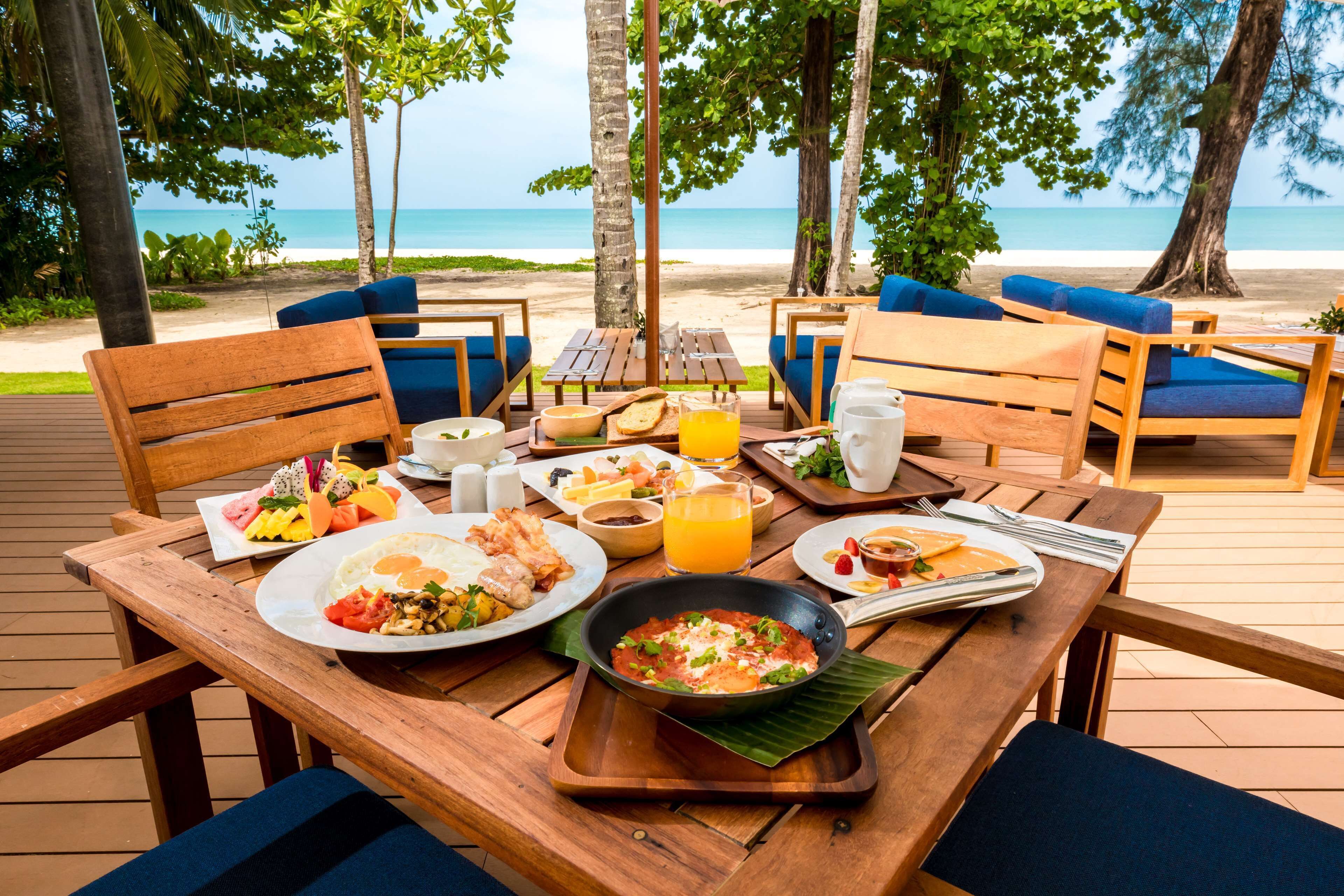 A wooden table set with breakfast items, including eggs, toast, and fruit, overlooking a beach.