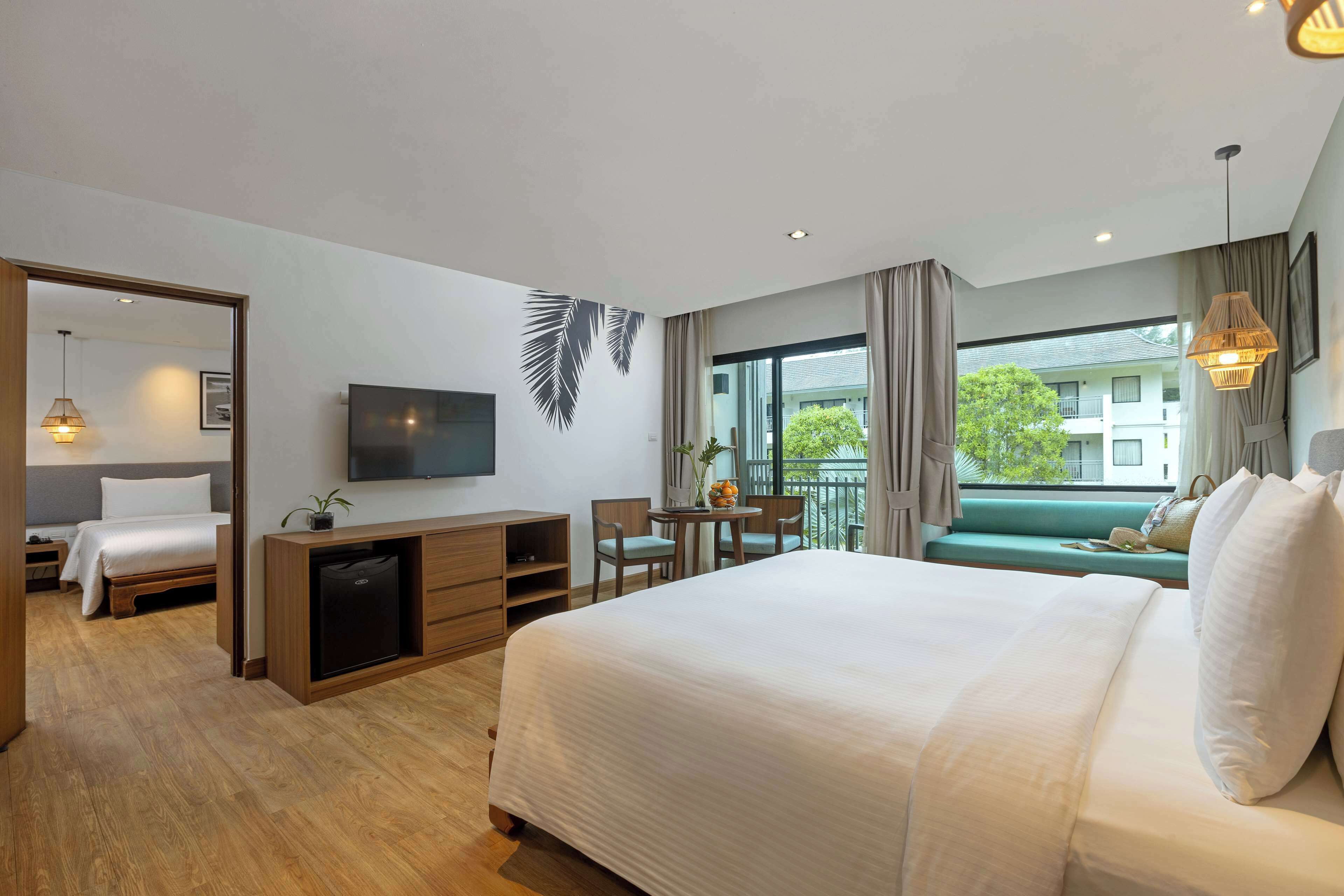 A modern hotel room with a large bed, a desk with a chair, a television, and a balcony with a view of buildings.