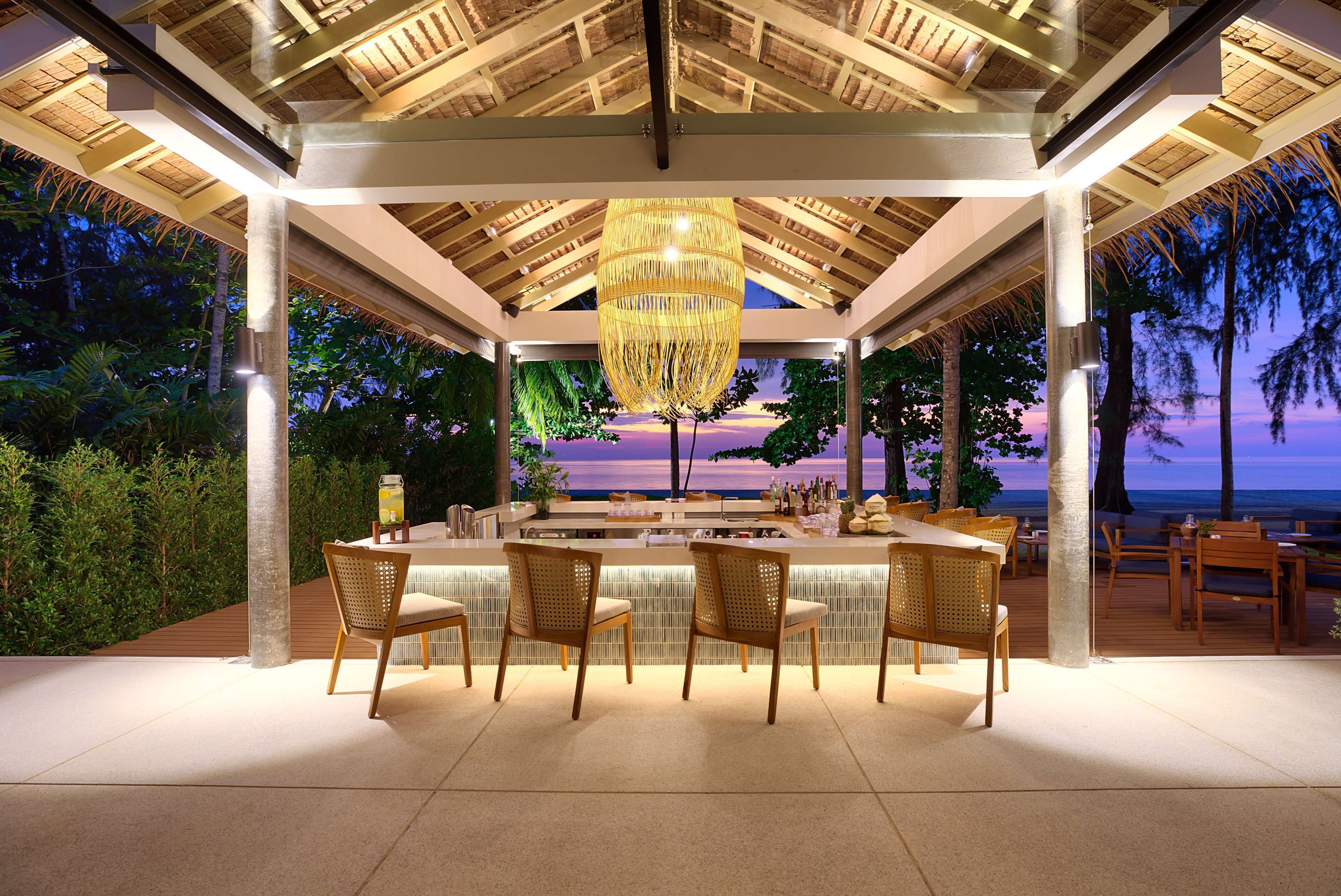 An outdoor bar area with a view of the ocean, wooden chairs, and a hanging light fixture.