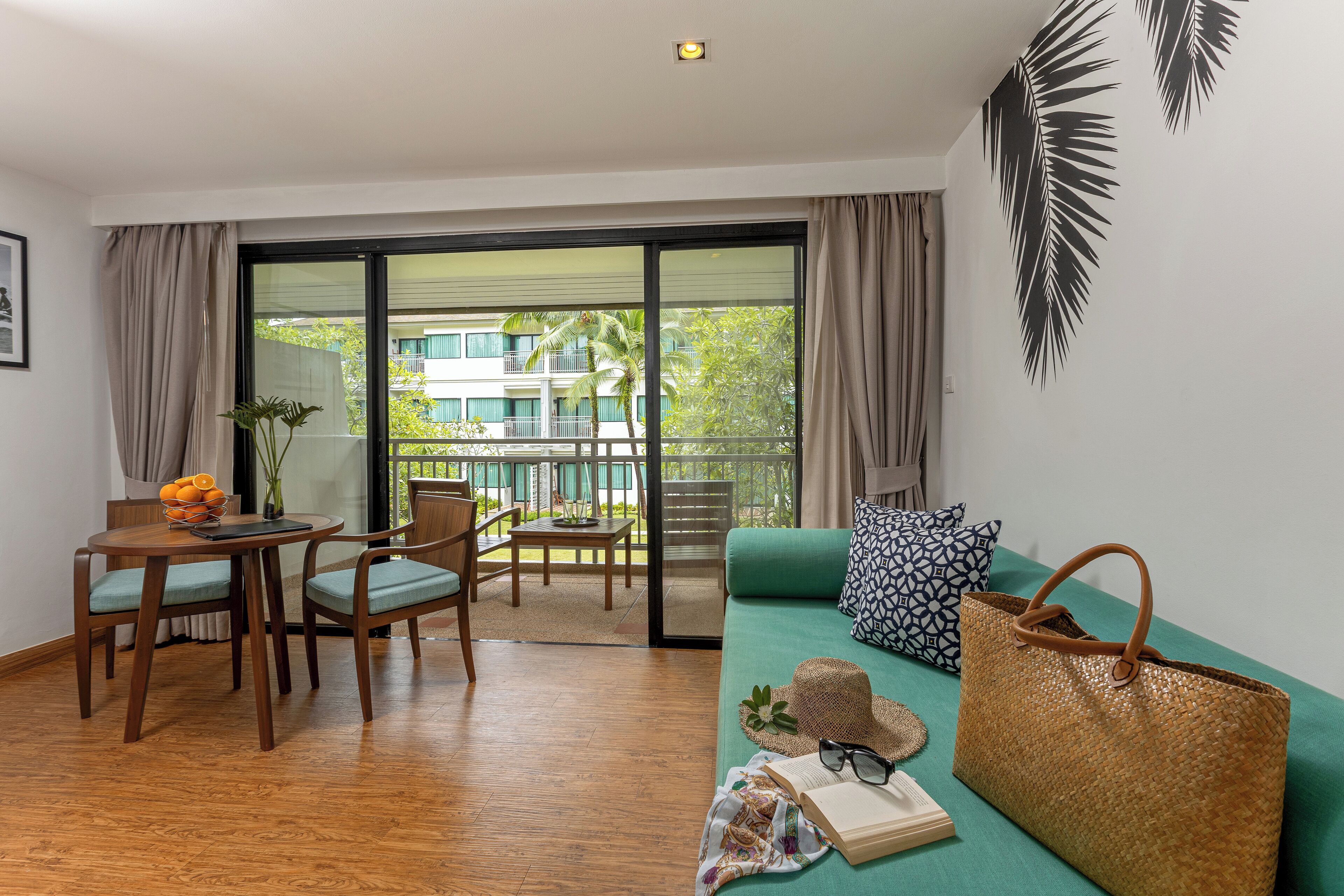 Una habitación con piso de madera, un sofá verde, una mesa de comedor con sillas, un balcón con vistas a edificios y árboles, y un bolso tejido grande.