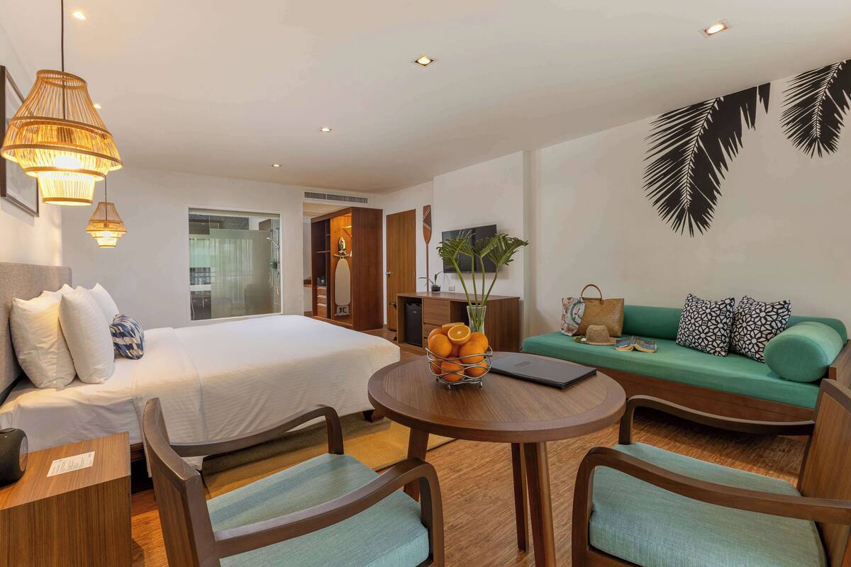 Une chambre d’hôtel comprenant un lit, un canapé, une table ronde avec des chaises, un téléviseur fixé au mur et une plante.