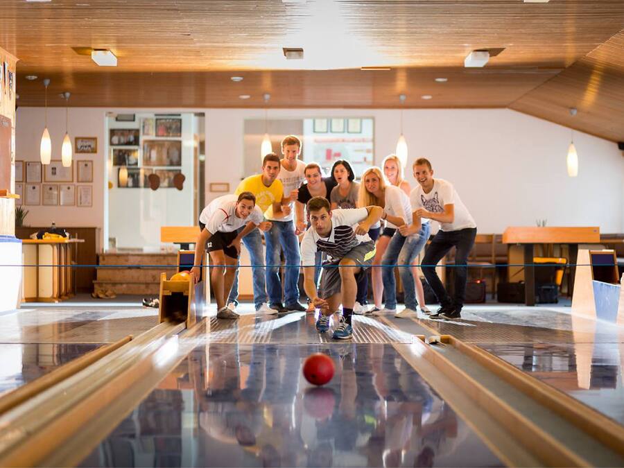 A bowling alley with a group of people gathered around a bowling lane, a red bowling ball, and wooden tables and chairs in the background.