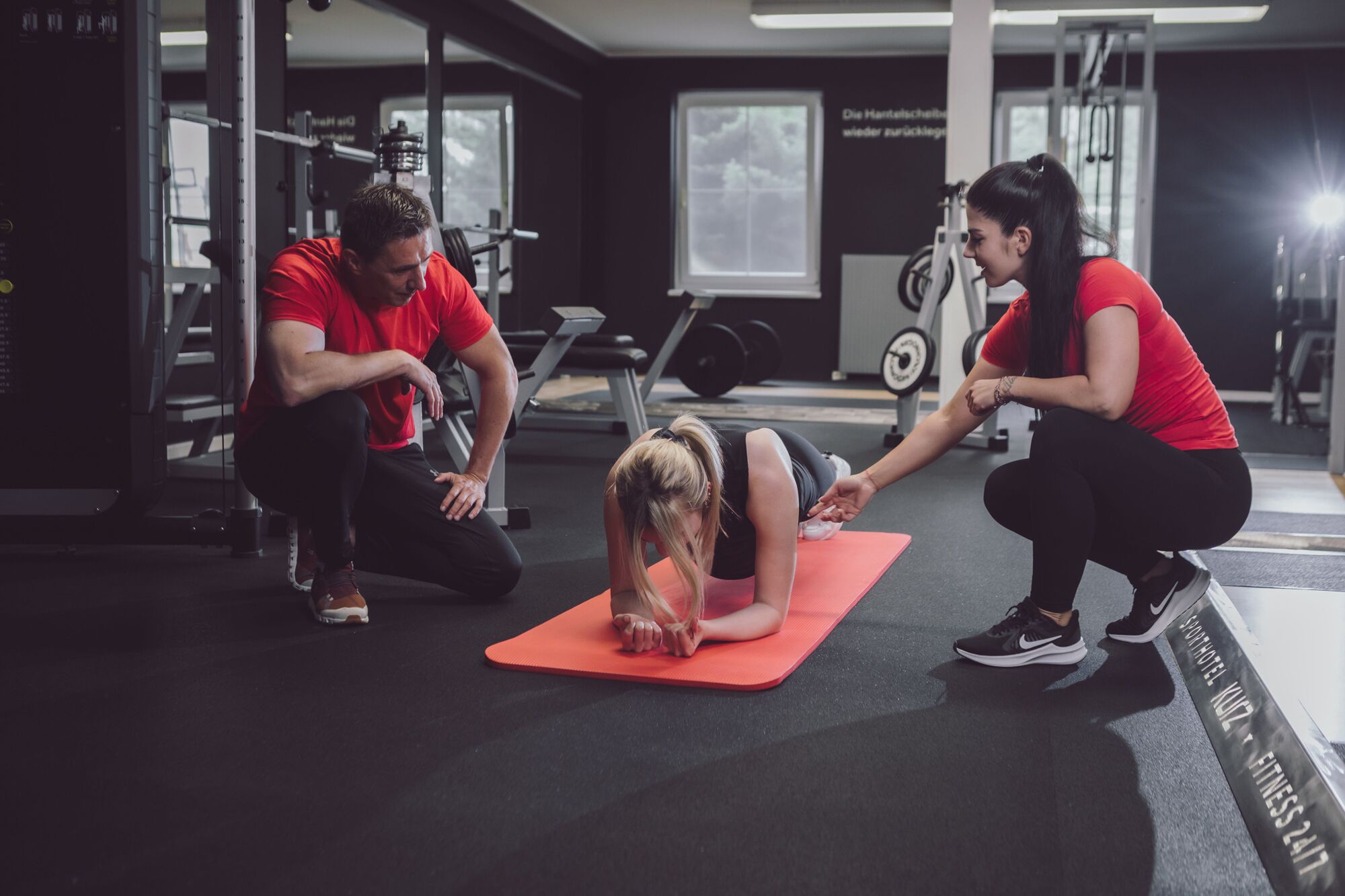 A gym with exercise equipment, a person doing a yoga pose on a mat, and two others observing.