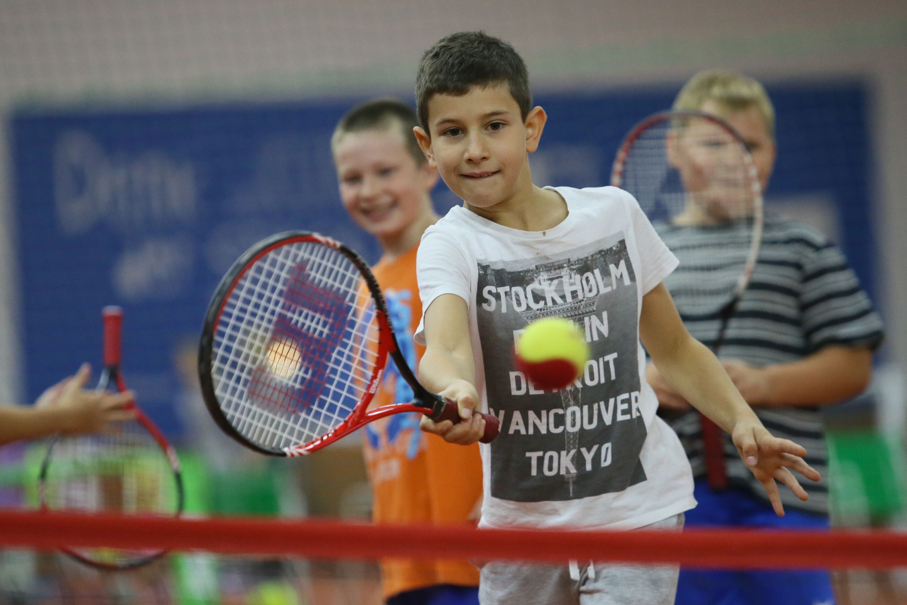 Un niño sosteniendo una raqueta de tenis, con otro niño al fondo.