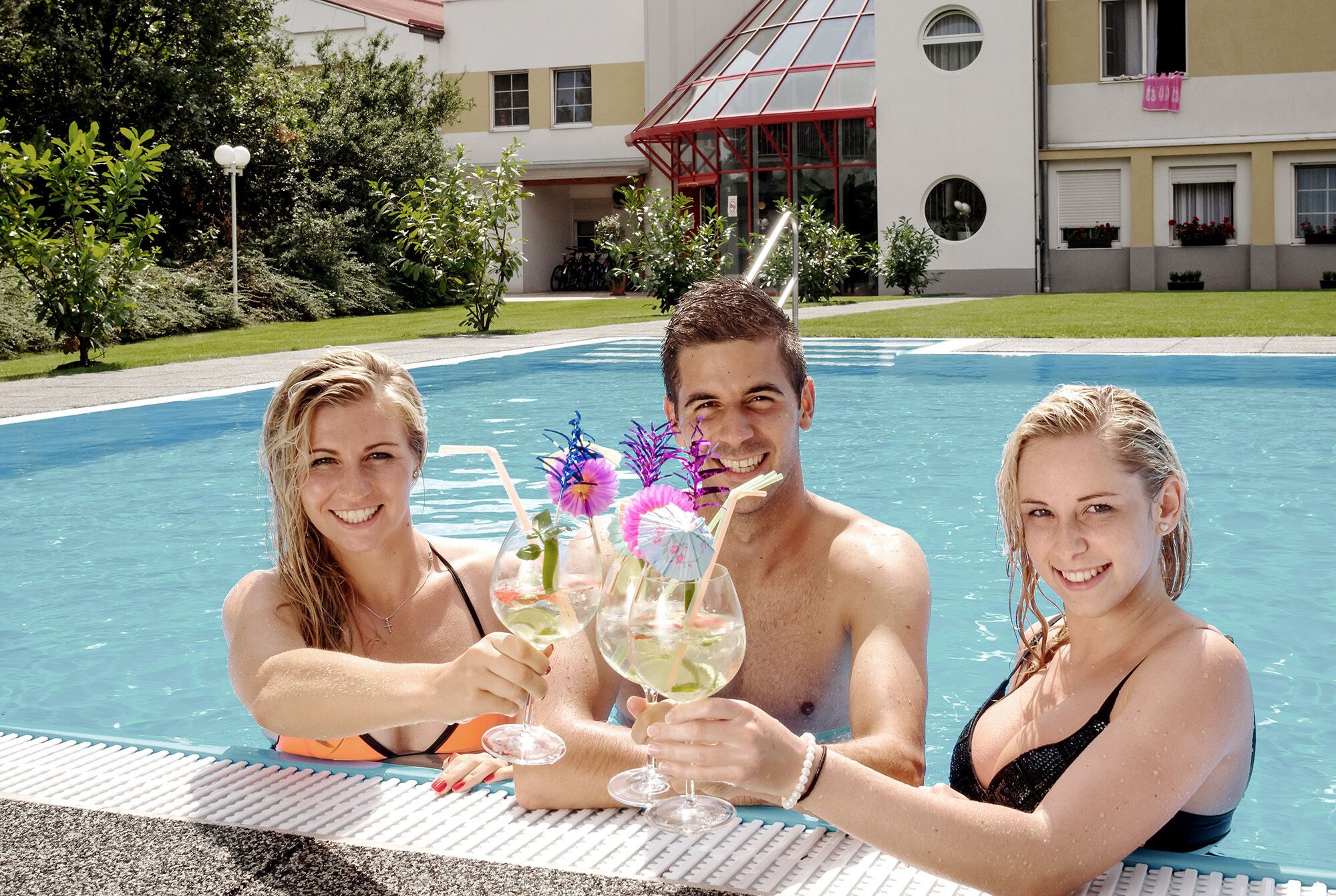 Ein Schwimmbecken im Vordergrund mit Tisch und Stühlen, umgeben von einem Gebäude mit rotem Dach und Fenstern.