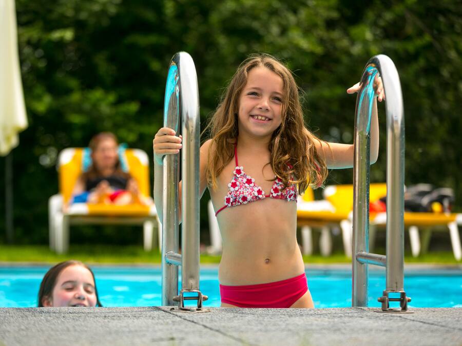 Un área junto a la piscina con un niño de pie en el borde, agarrado a escaleras de metal. Se observan sillas de descanso y sombrillas al fondo.
