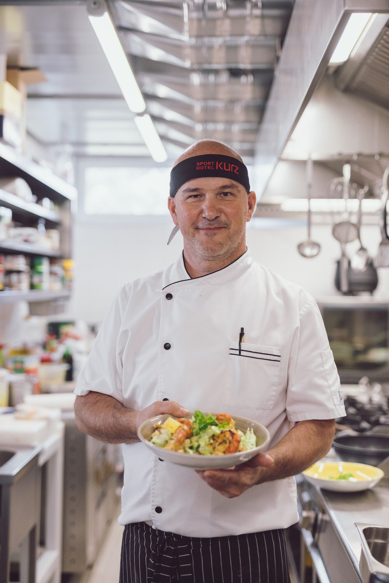 A chef in a white uniform holding a plate of food in a kitchen.