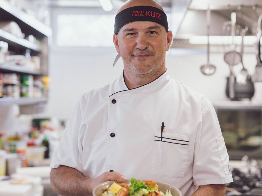 A chef in a white uniform holding a plate of food in a kitchen.