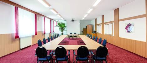A conference room with a long table, blue chairs, a projector screen, and a potted plant.