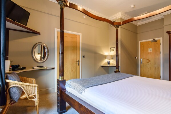 A bedroom with a four-poster bed, a wicker chair, a bedside table with a lamp, and a mirror on the wall.