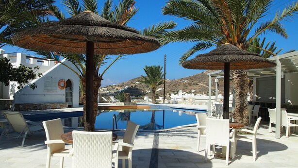 A poolside area with white wicker chairs and thatched umbrellas, surrounded by palm trees and a clear blue sky. - Homer's Inn Hotel (Ios)