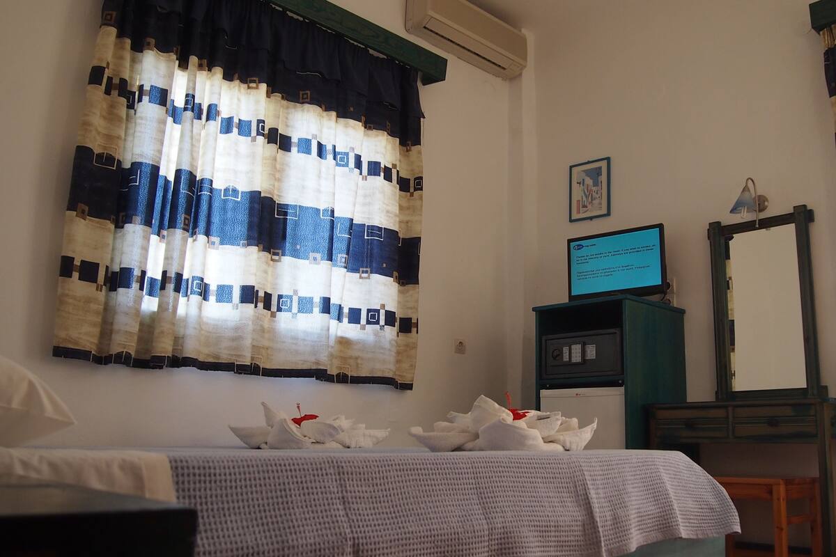 Hotellihuone, jossa on raidalliset verhot, ruutukuviollinen peitto sängyllä, työpöytä, jossa on tietokone, peili ja seinälle asennettu ilmastointilaite.