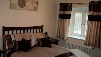 Ein ordentlich bezogenes Bett mit einem Holz-Kopfteil, ein Nachttisch mit Lampe und ein Fenster mit Vorhängen.