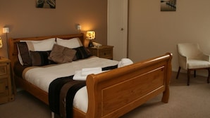 A bedroom with a wooden bed, bedside tables, a chair, and two wall-mounted lamps. - The Royal Lodge (Ross-on-Wye)