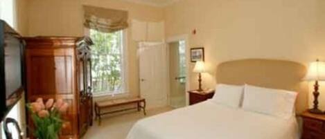 A bedroom with a large bed, a bedside table with a lamp, a wooden wardrobe, and a window with curtains.