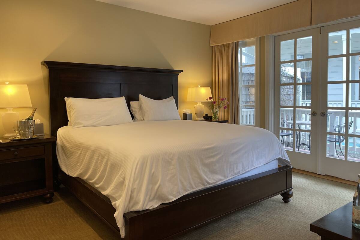 A neatly made bed with white linens in a hotel room with a dark wooden headboard, two bedside lamps, and a view of the outside through a large window.
