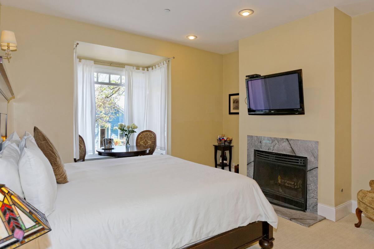 A bedroom with a large bed, a television mounted on the wall, a fireplace, and a window with curtains.