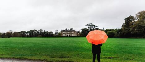 Une personne se tient sur une route mouillée, un parapluie orange à la main, et fait face à un grand bâtiment situé au loin.