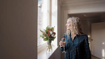 A person holding a glass with a drink, standing near a window with a vase of flowers.