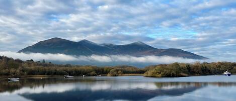 A calm lake with boats, surrounded by trees and mountains under a cloudy sky.