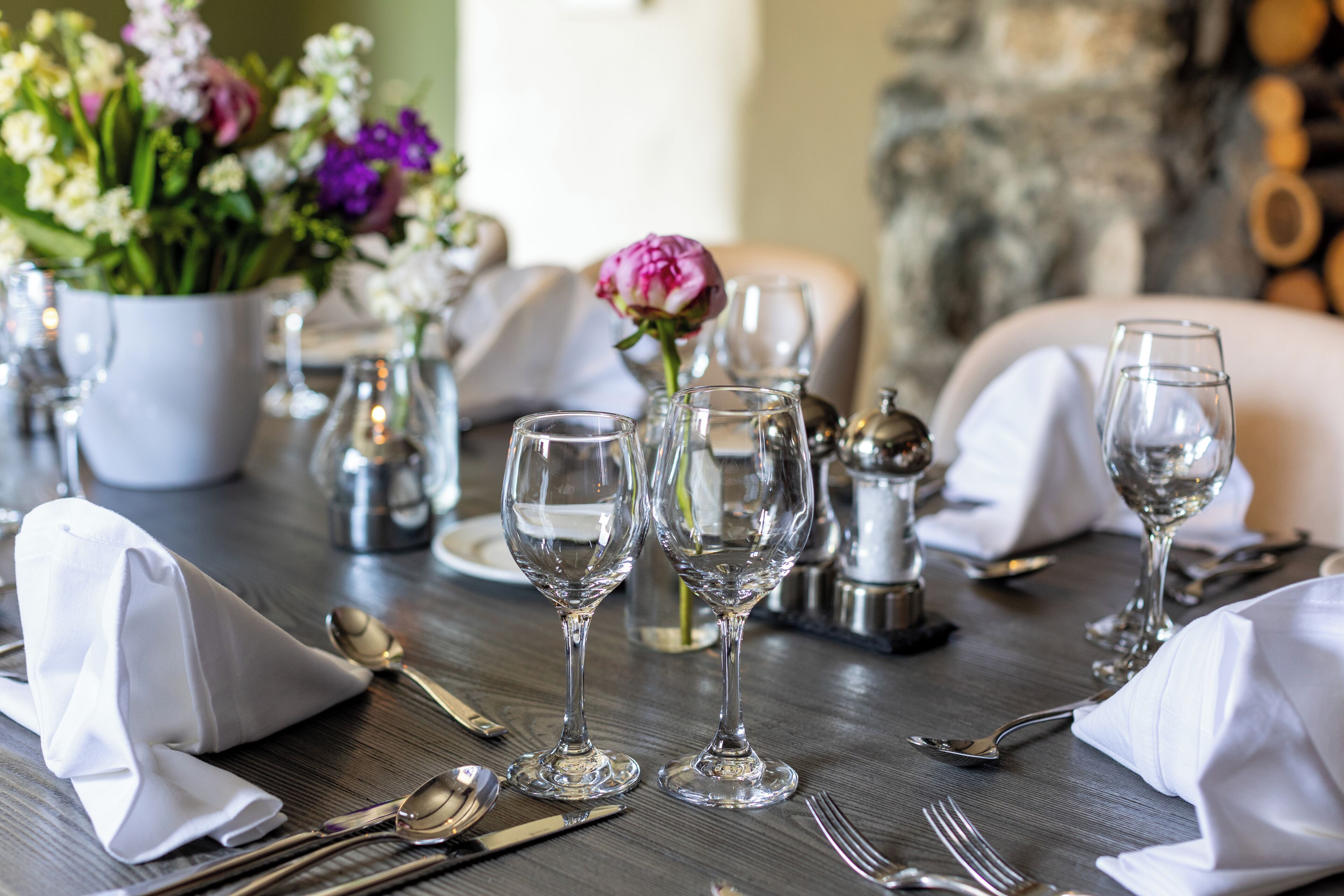 A dining table set with white napkins, silverware, and wine glasses, with a vase of flowers in the background.