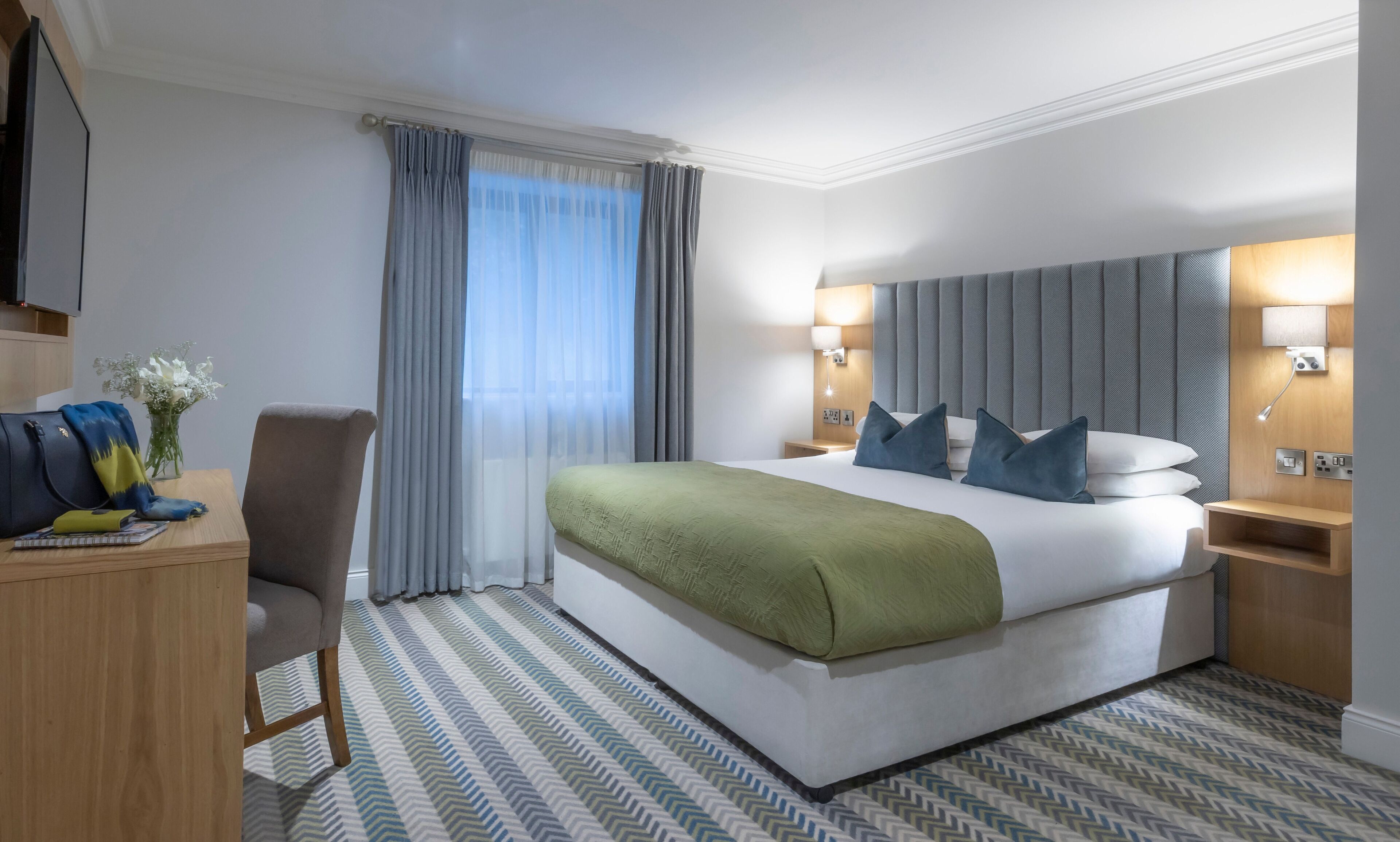 Quarto de hotel com uma cama grande, uma mesa, uma cadeira, mesa de cabeceira e luminária na parede.