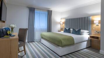 A hotel room with a large bed, a desk, a chair, a nightstand, and a wall-mounted lamp.