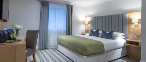 Quarto de hotel com uma cama grande, uma mesa, uma cadeira, mesa de cabeceira e luminária na parede.