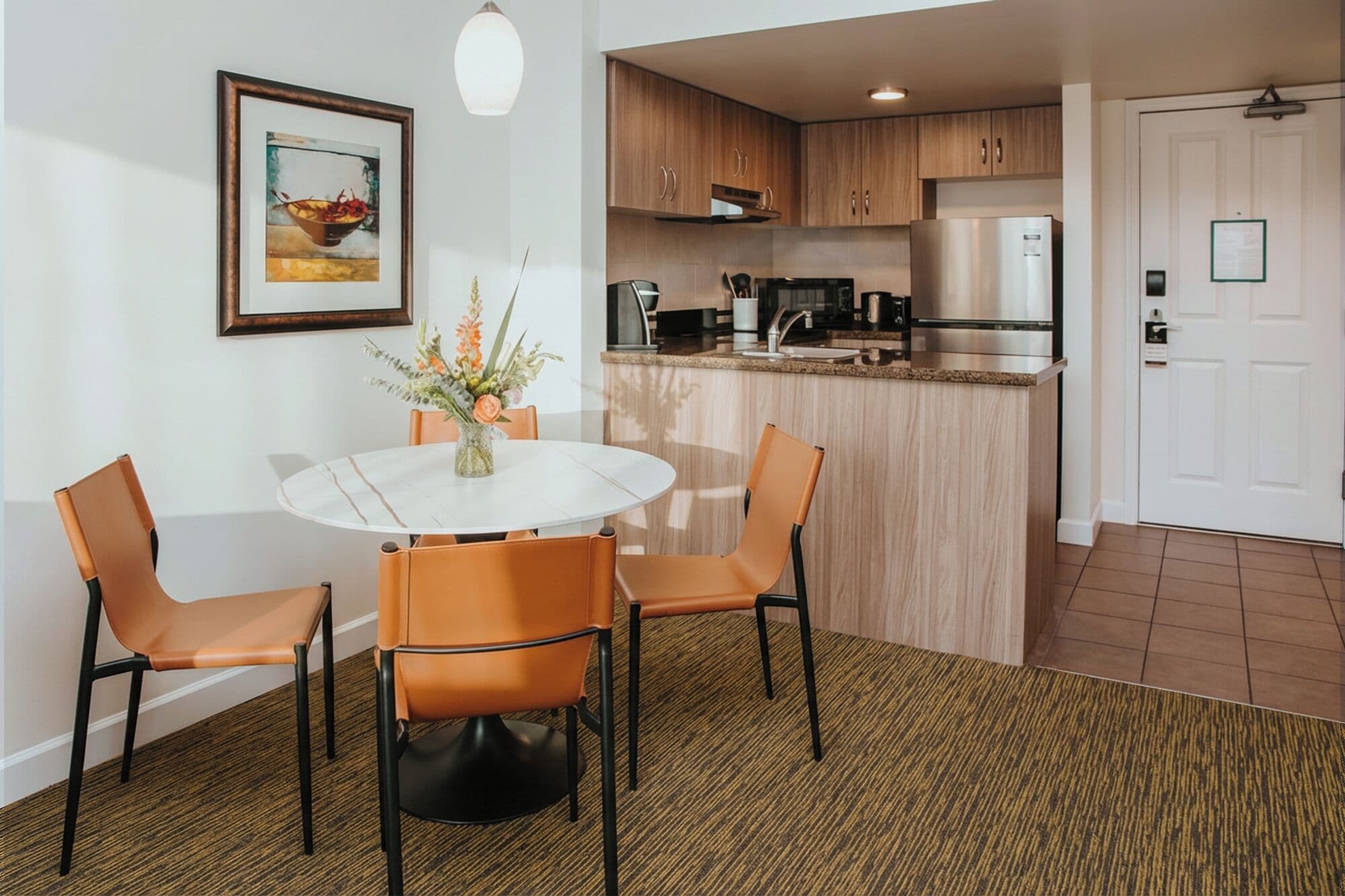 A dining area with a round table, four chairs, a vase with flowers, and a kitchen in the background.