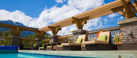 A poolside area with wooden pergolas, lounge chairs, and a view of mountains.
