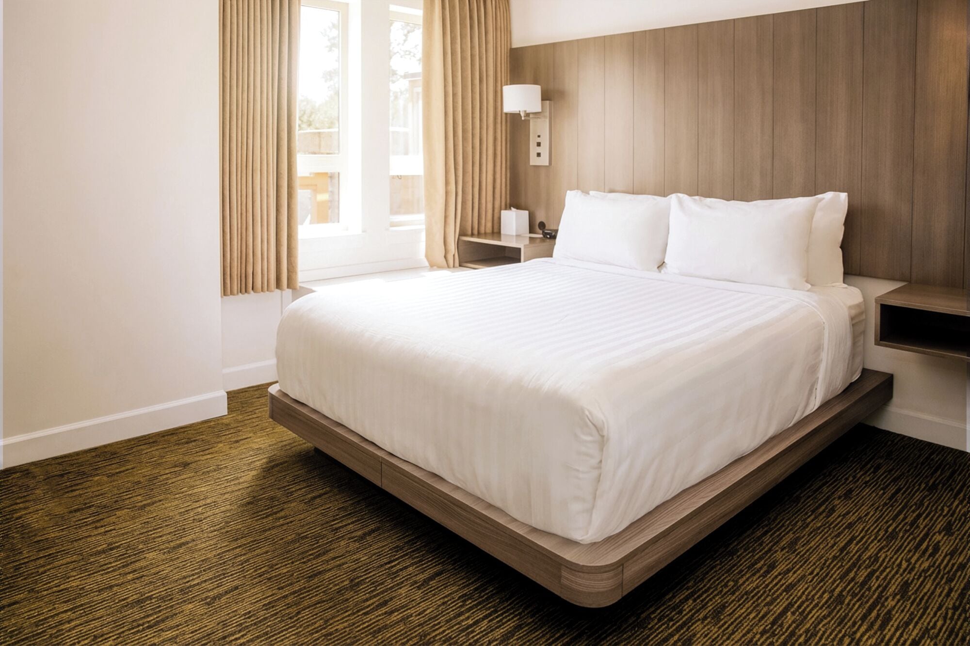 A hotel room with a large bed, wooden headboard, a nightstand, and a window with curtains.