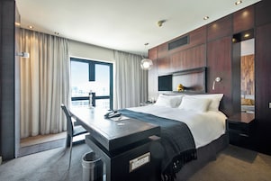 A modern hotel room with a large bed, a desk, a chair, a desk lamp, a nightstand, and a view of the city.