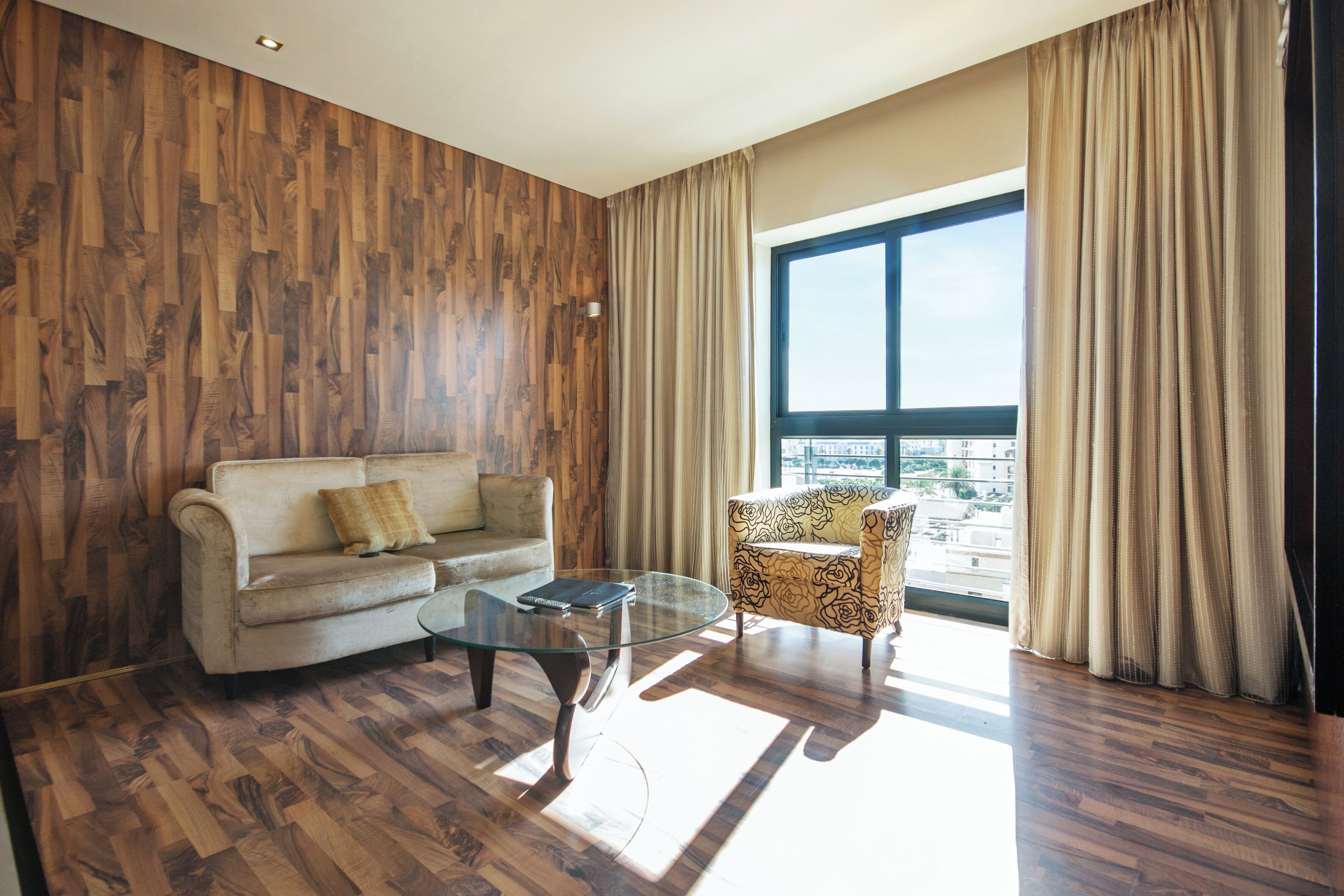 Sala de estar con sofá, sillón, mesa de centro de vidrio, piso de madera y un ventanal con cortinas.