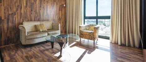 Sala de estar con sofá, sillón, mesa de centro de vidrio, piso de madera y un ventanal con cortinas.