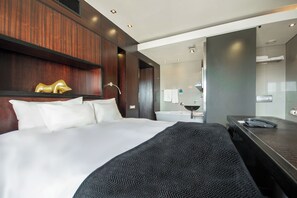 A hotel room with a large bed, a bedside table, a mirror, a bathroom with a sink and toilet, and a wooden accent wall. - Colosseum Hotel (Cape Town)