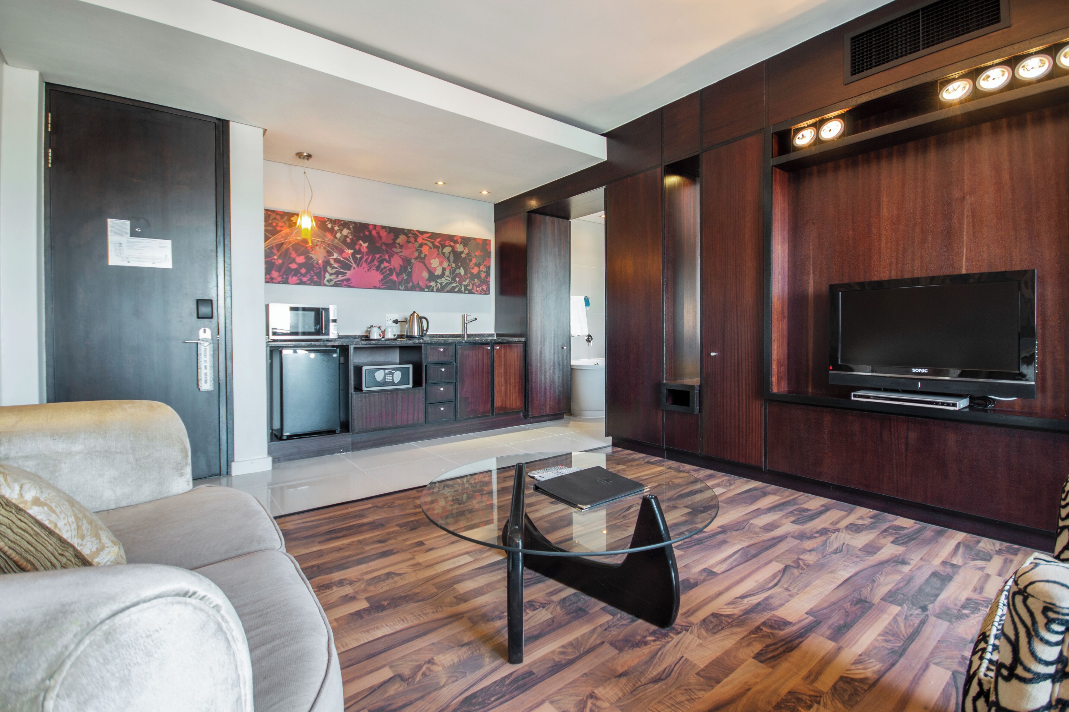 A modern living room with a sofa, a glass coffee table, a flat-screen TV, and a kitchen in the background.